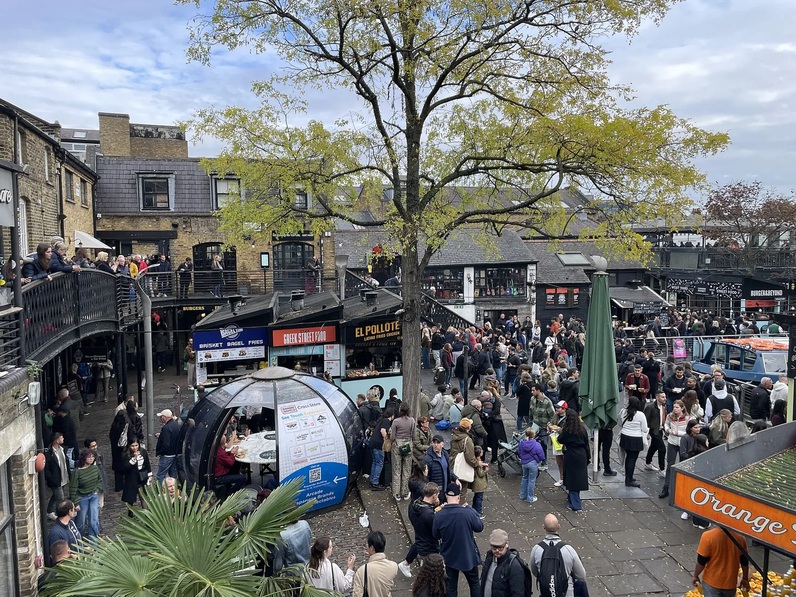 The yard of Camden Lock Market full of people among food stalls, with a tree in the middle and brick warehouses behind