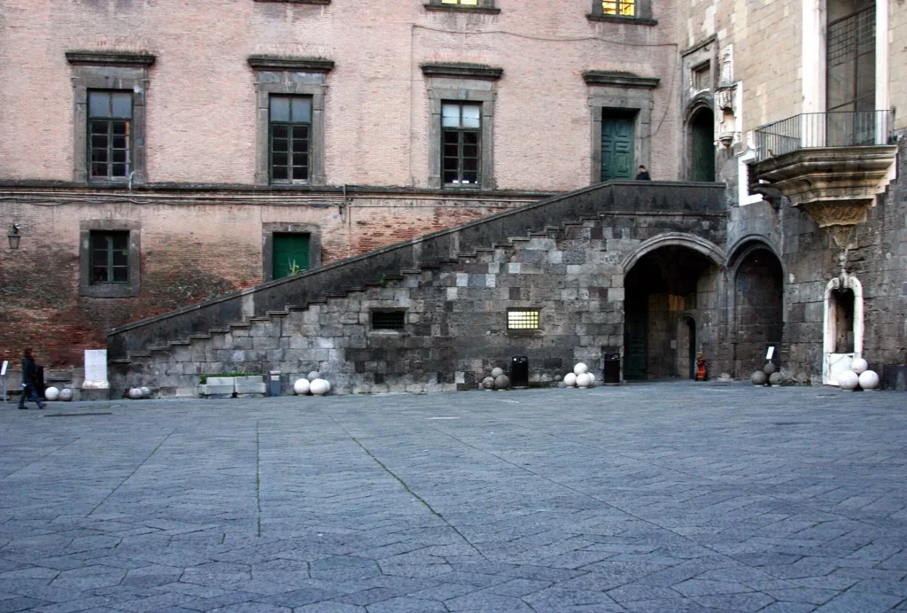 Courtyard of Castel Nuovo: an outdoor staircase of grey stone climbing along a pink brick wall to a doorway, with low arches at its foot and stone cannonballs on the paving
