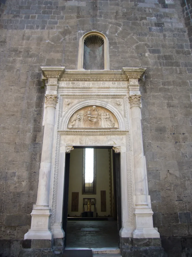White marble Renaissance doorway of the Palatine Chapel in a wall of dark stone blocks: fluted columns with Corinthian capitals, a carved relief in the lunette above the door and an empty niche at the top