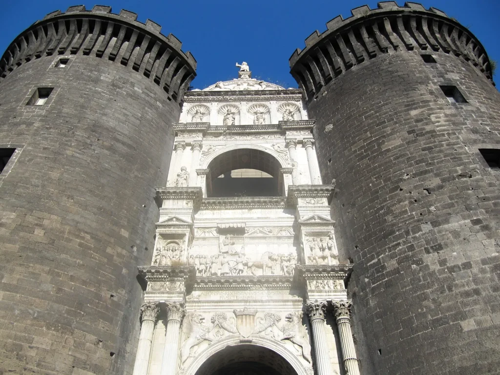 The white marble triumphal arch of Castel Nuovo seen from below, set between 2 dark round towers: paired columns, a carved frieze and an upper arch, with a statue on top