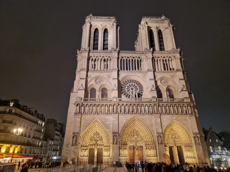 The west front of Notre-Dame de Paris lit up at night, with its two towers, central rose window and three portals, and people on the square