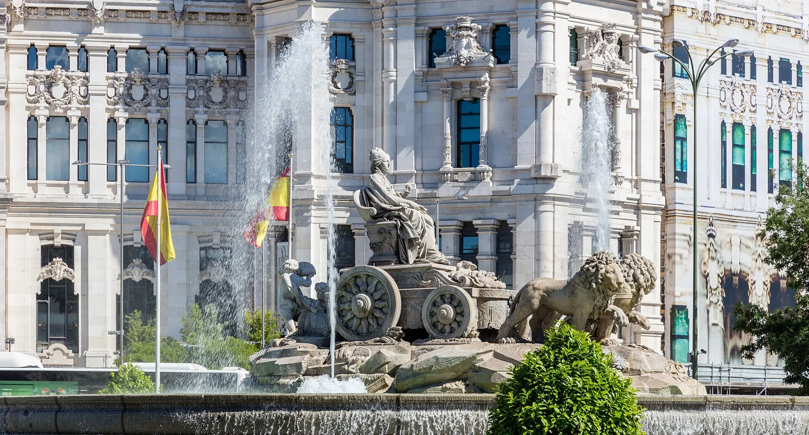 The Cibeles Fountain, with the goddess in her chariot drawn by 2 lions and a tall water jet, in front of the white facade of the Palacio de Cibeles with Spanish flags