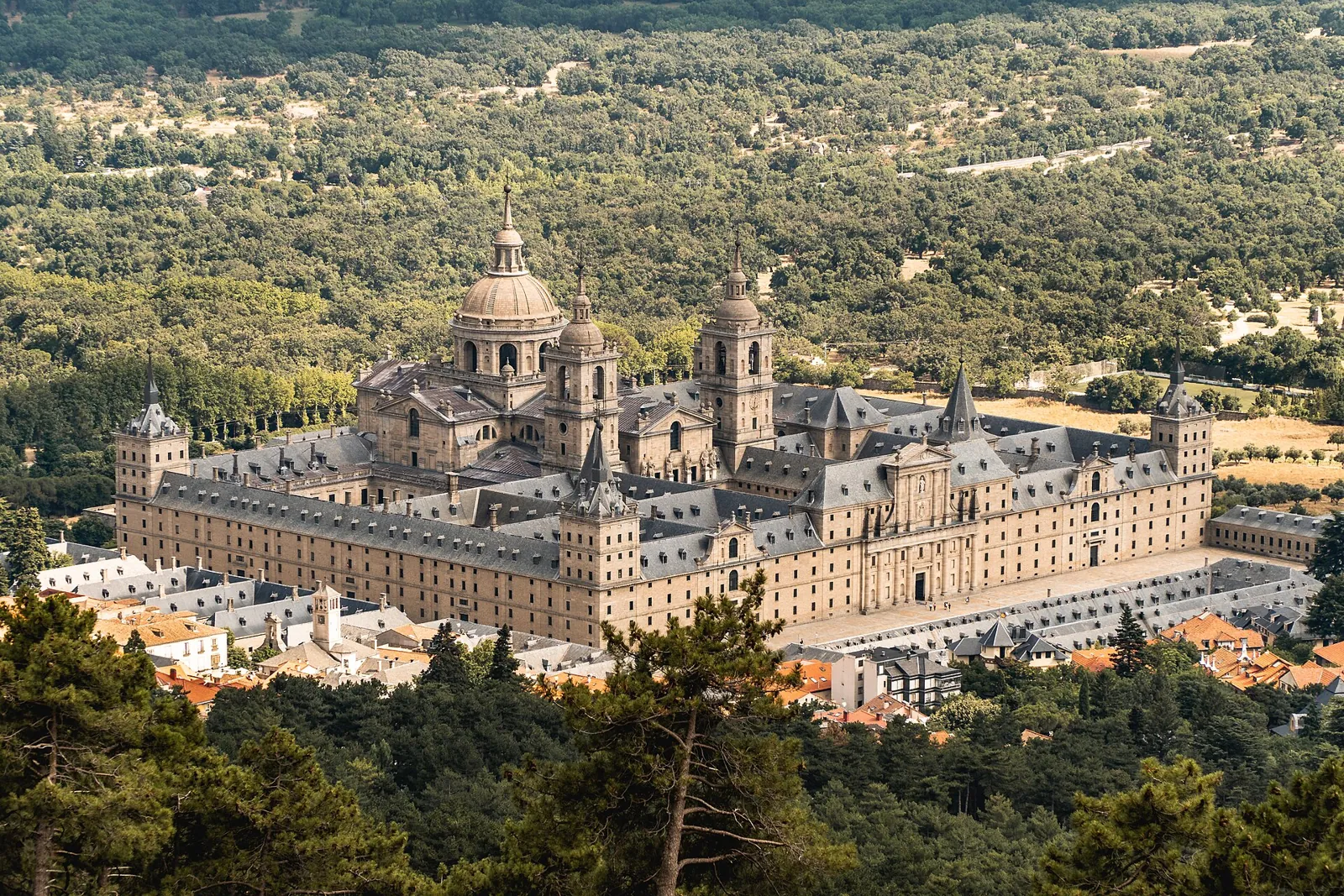 Overall view of the Royal Monastery of El Escorial, a large granite building with a dome and towers, surrounded by trees and the rooftops of the town
