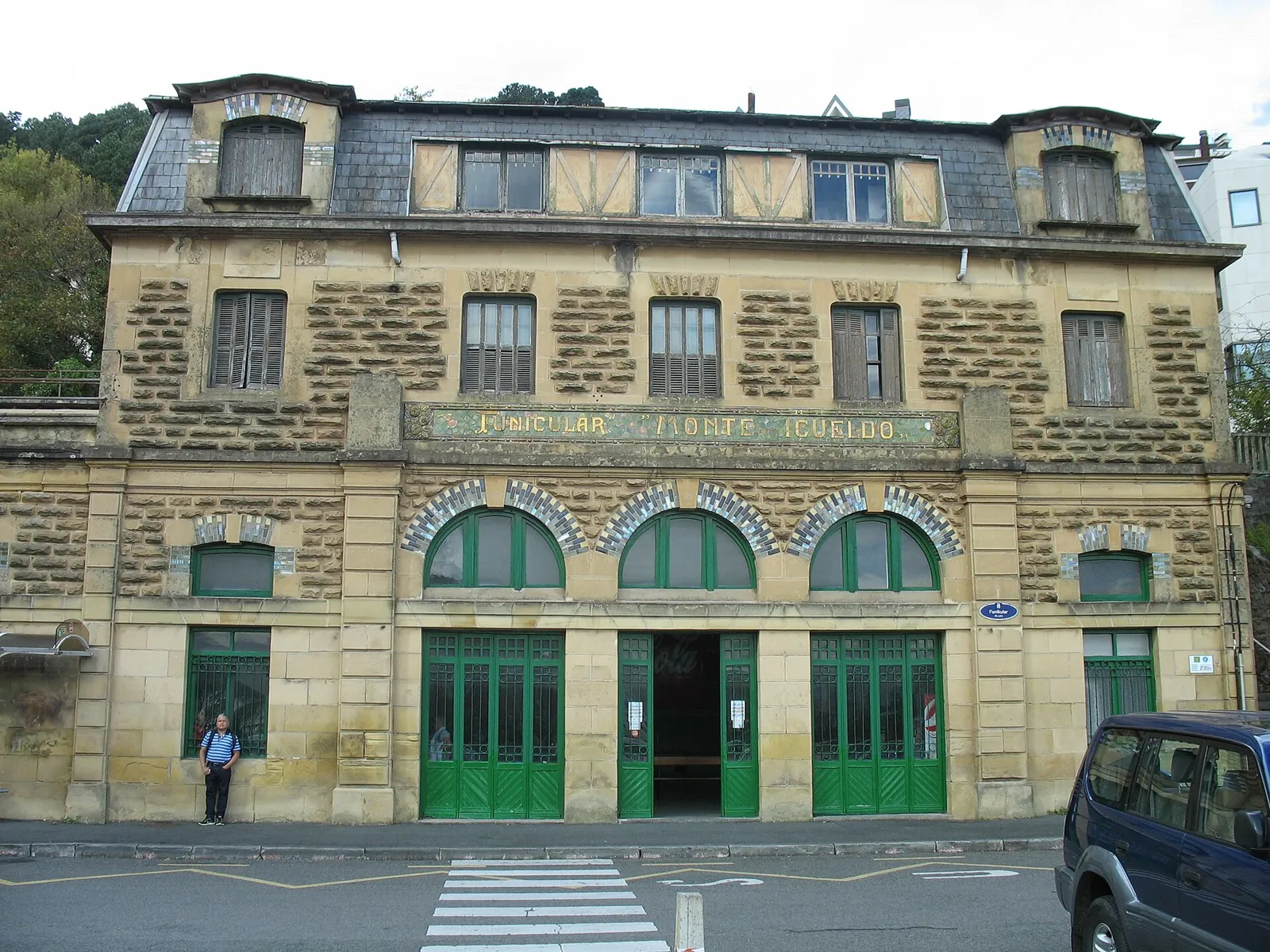 Stone façade of the lower station of the Igueldo funicular, with a slate roof, 3 arches over green doors and the funicular sign