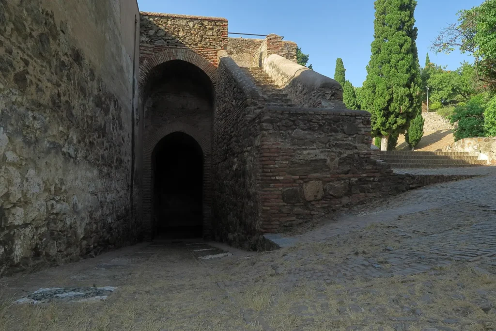 Arched entrance through a stone and brick tower at Gibralfaro Castle, with a cobbled ramp and cypress trees behind