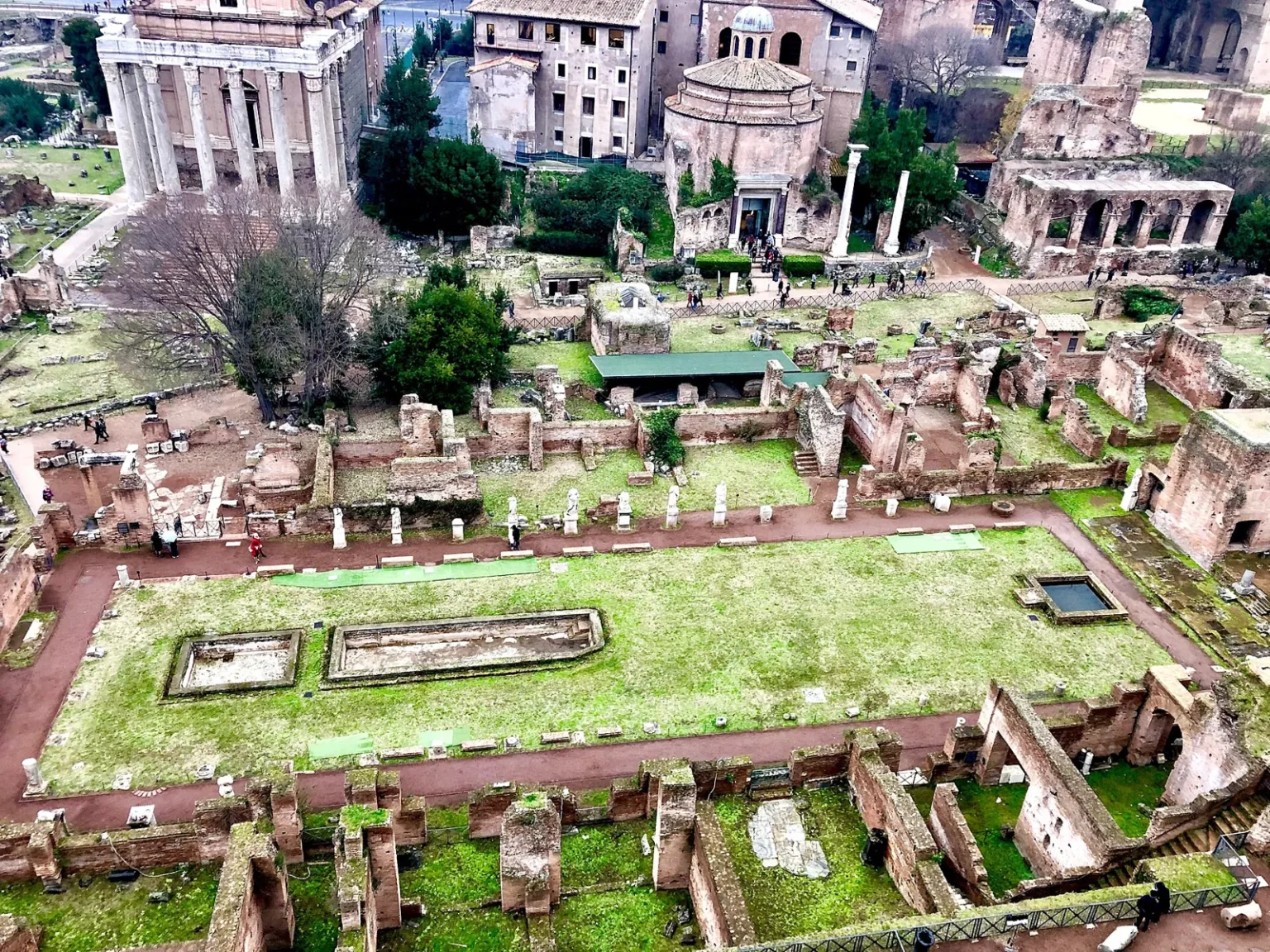 The House of the Vestals seen from the Palatine Hill: a long lawn with rectangular pools framed by brick walls and a row of white statues on pedestals, with the round Temple of Romulus and the columns of the Temple of Antoninus and Faustina behind