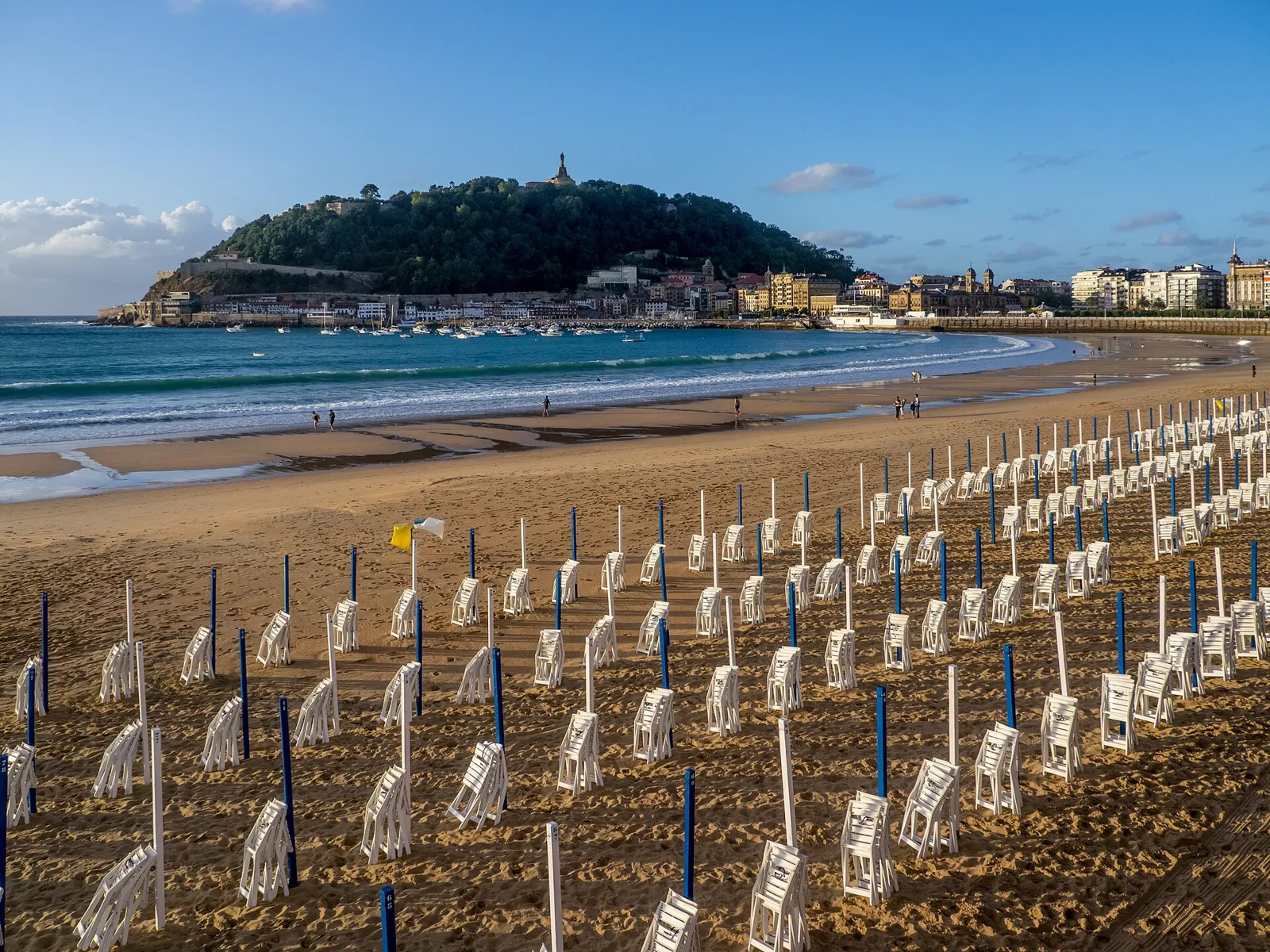 Rows of blue-and-white sunshade poles, without their canvas, and folded white chairs on the sand of La Concha at dusk, with Mount Urgull in the background