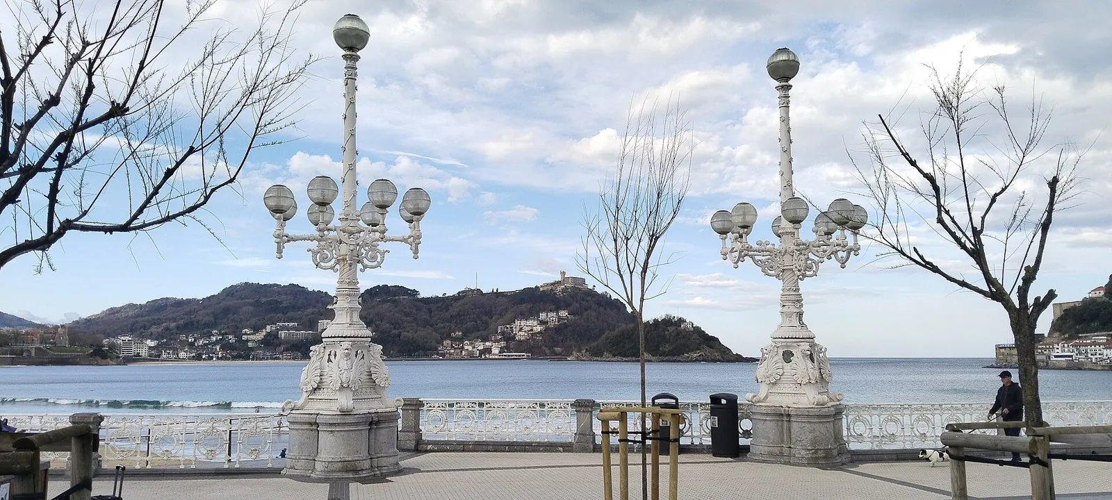 Two white multi-armed lamp posts and the white railing of the La Concha promenade, with the bay and Mount Igeldo behind