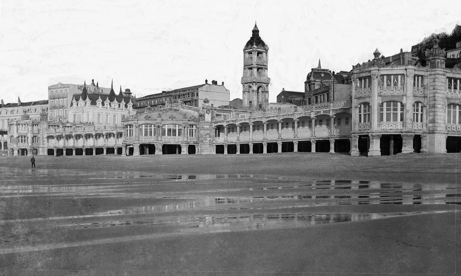 Old black-and-white photograph of the La Perla del Océano bathhouse on La Concha beach, with wet sand in the foreground