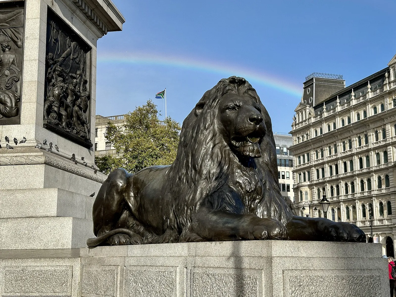 One of Landseer's 4 bronze lions lying at the foot of Nelson's Column, next to a bronze relief, with a rainbow in the sky