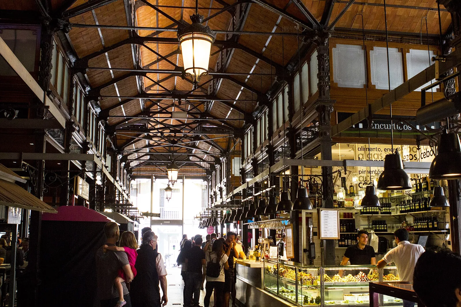 Inside the Mercado de San Miguel: a single open hall with iron columns and roof trusses, hanging lanterns and counters along the sides