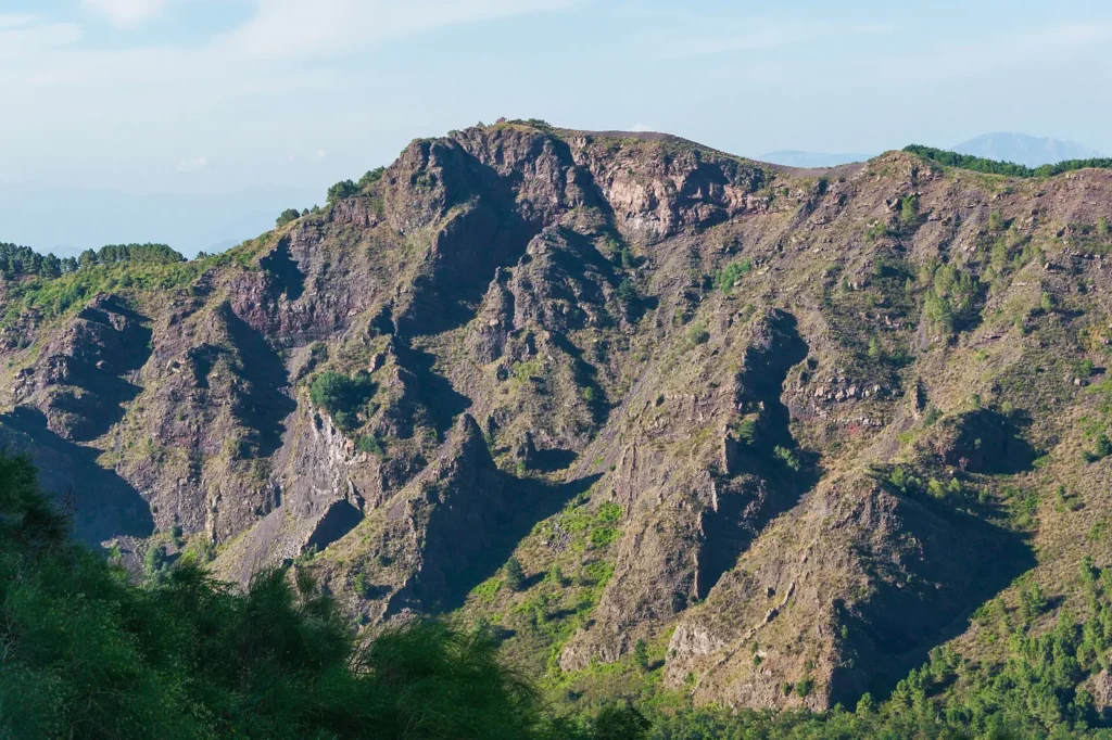 The steep inner wall of Monte Somma seen from Vesuvius: jagged ridges of reddish and grey volcanic rock cut by gullies, with pine trees and shrubs on the lower slopes