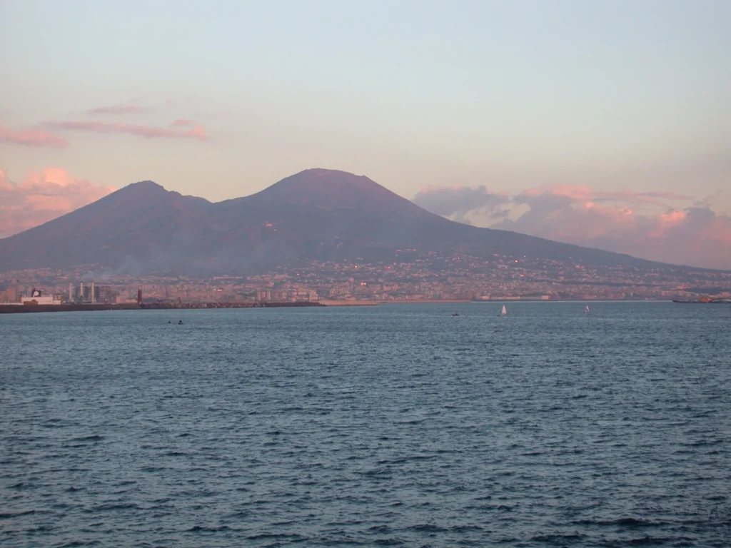 Mount Vesuvius seen across the Bay of Naples at sunset: the lower, jagged ridge of Monte Somma on the left and the higher, rounded Great Cone on the right, above a band of houses and a harbour breakwater
