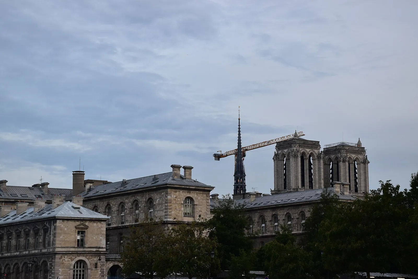 The towers and spire of Notre-Dame behind the rooftops of the Île de la Cité, with a construction crane beside the spire on a cloudy day