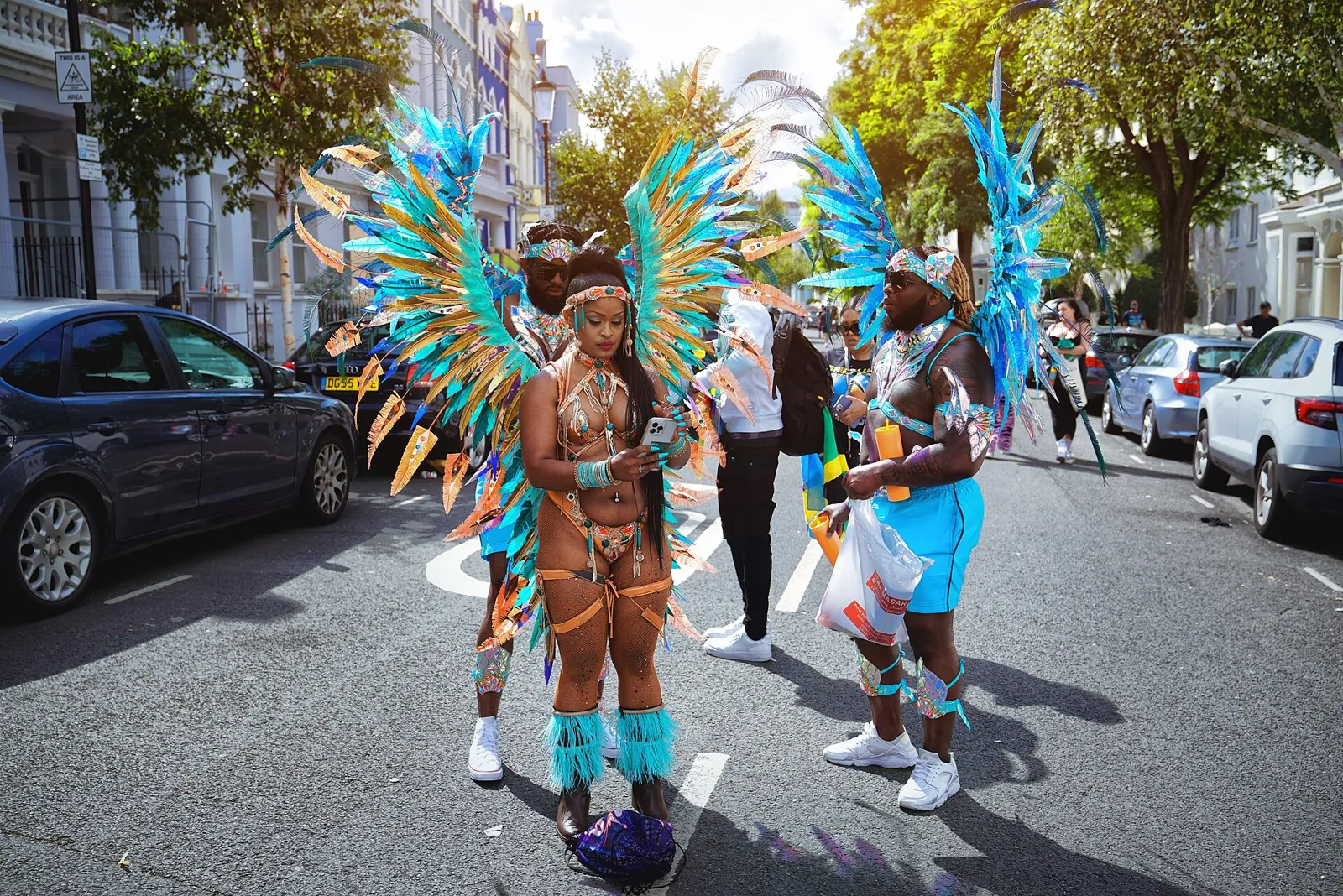 Three members of a Notting Hill Carnival mas band, a woman and two men, in turquoise and orange feathered wings, standing on a street of white houses and trees