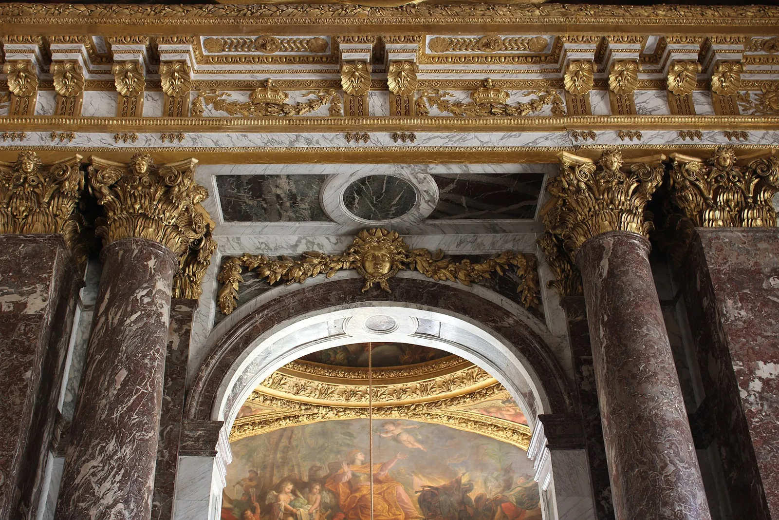 Red marble columns with gilded capitals and an arch beneath a painted ceiling in the Hall of Mirrors at Versailles