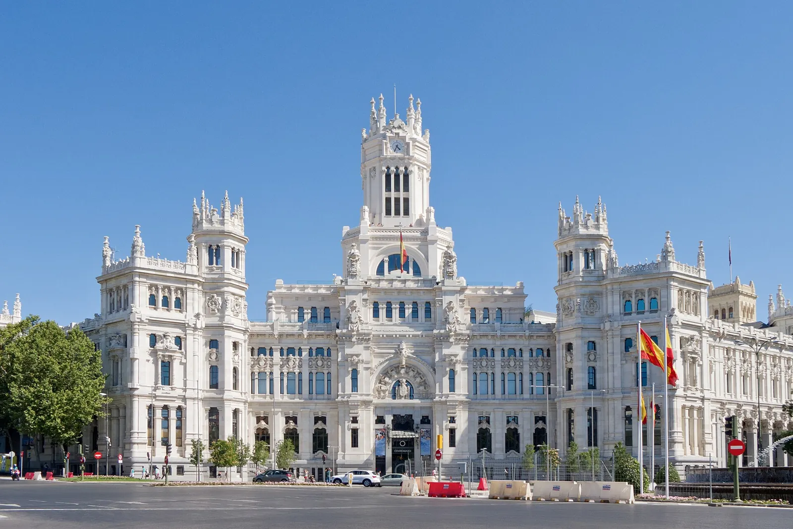 The white facade of the Palacio de Cibeles, with its central tower and 2 side towers topped with pinnacles, seen from the square on a clear day