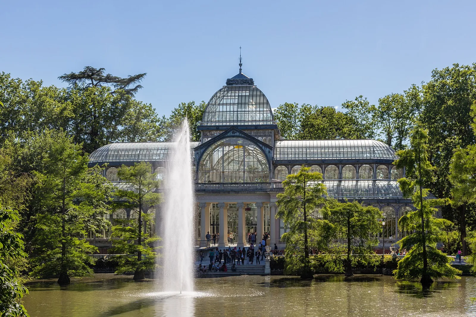 The Palacio de Cristal in Retiro Park seen from the front, with its iron and glass dome, the lake fountain in front and trees growing in the water