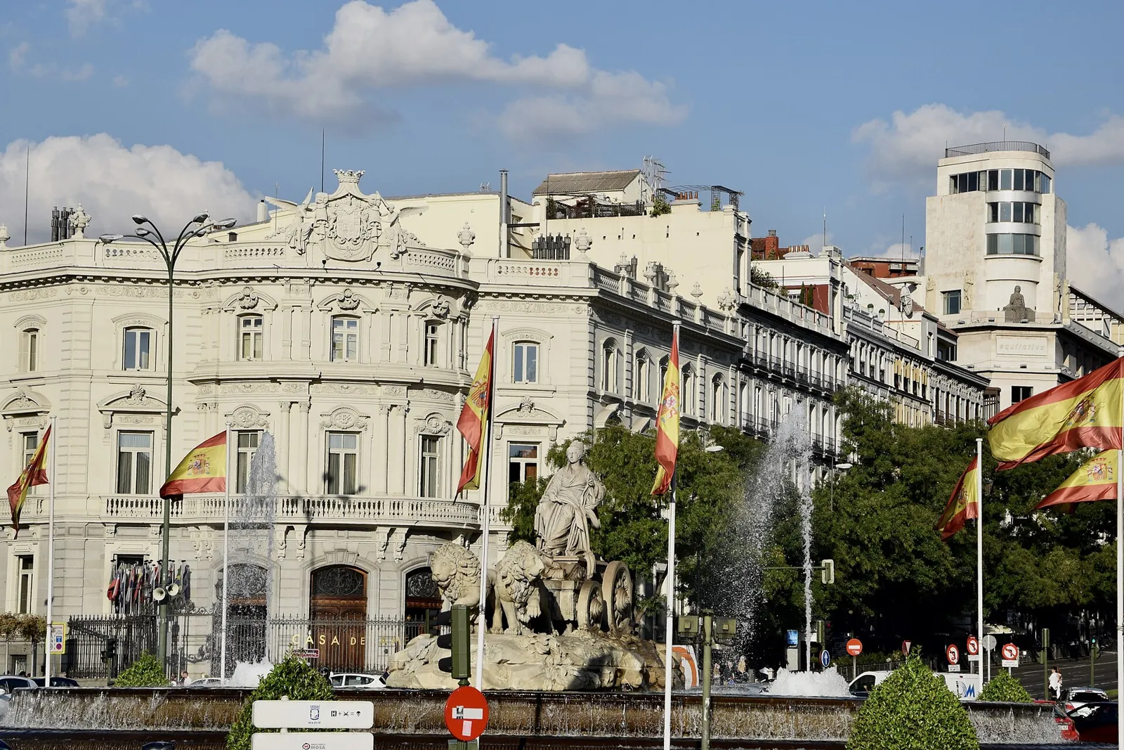 The Palacio de Linares, a pale building with balconies and Spanish flags in front, with the Cibeles Fountain and one of its water jets in the foreground