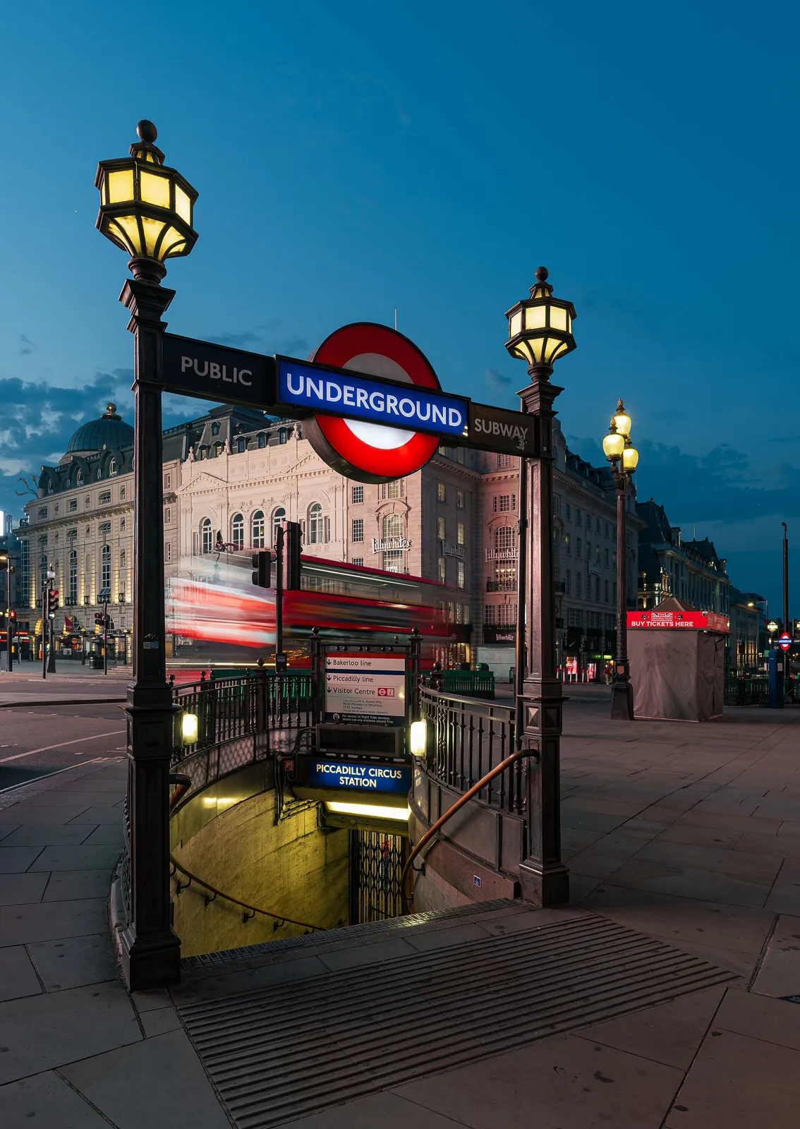 A Piccadilly Circus Tube entrance: a staircase going down under the Underground sign, between 2 lit lamps