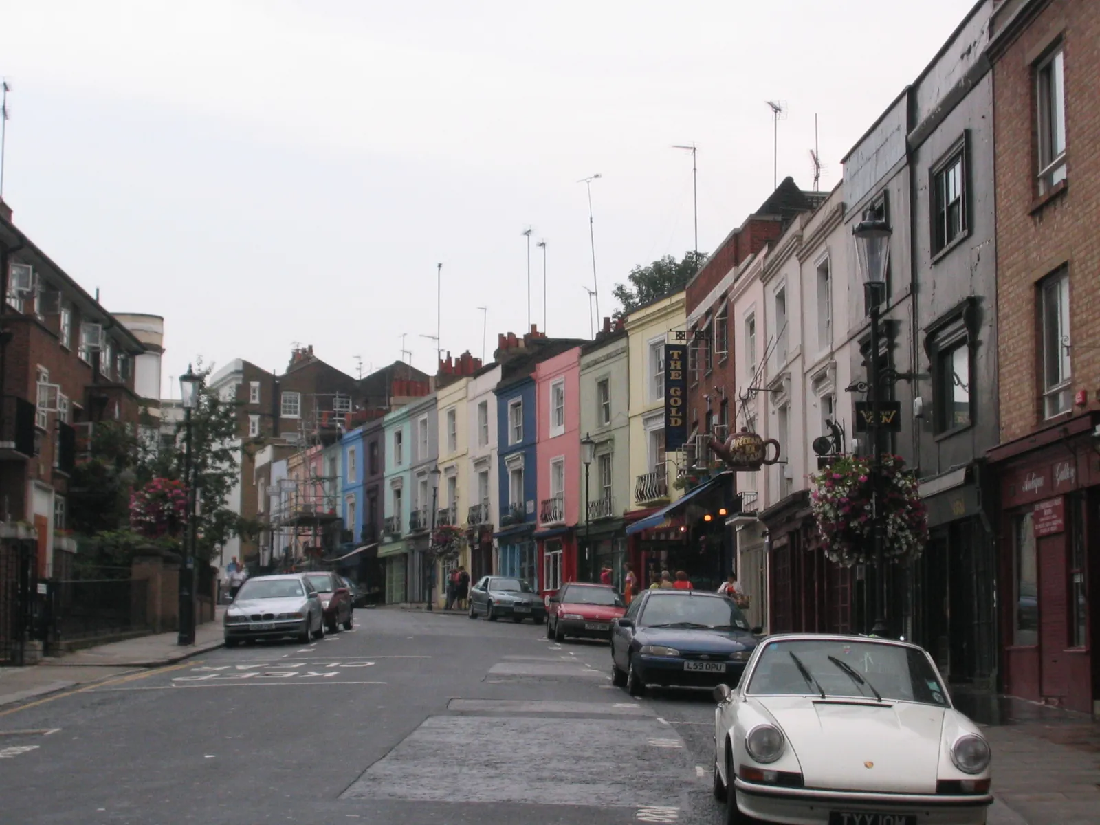 Portobello Road on a day without stalls, with houses painted blue, pink, green and yellow above the shops, flower baskets on the lamp posts and parked cars
