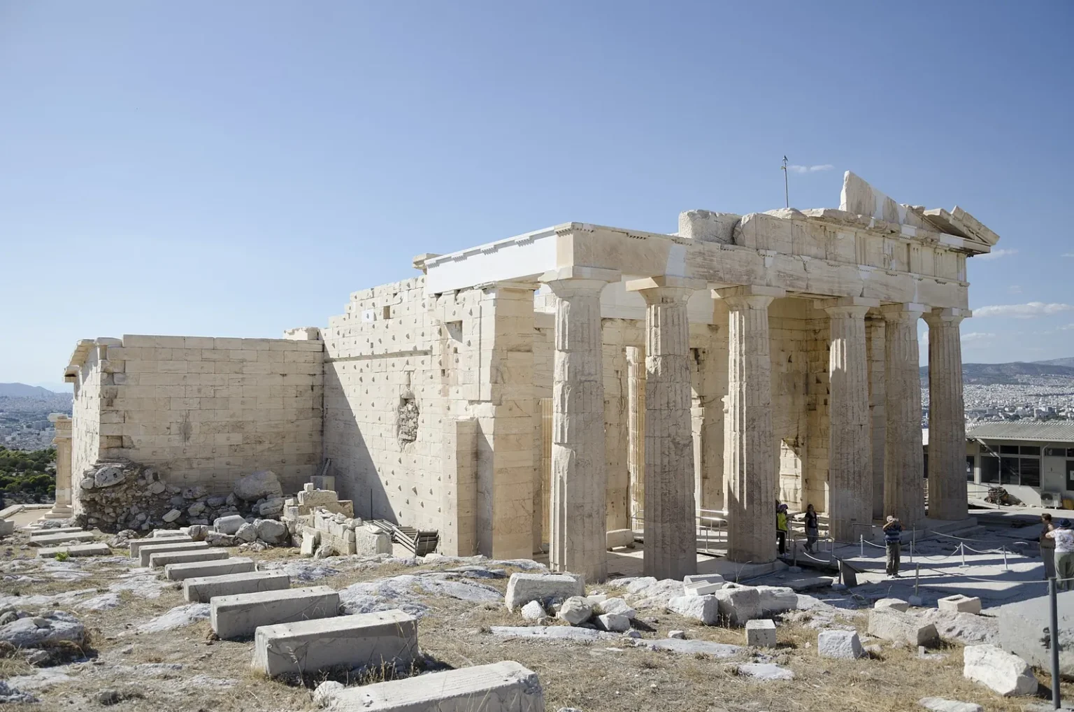 The Propylaea seen from the south-east, inside the Acropolis enclosure: the east porch with its Doric columns under the remains of the pediment, a tall wall of marble blocks on the left and a few visitors between the columns