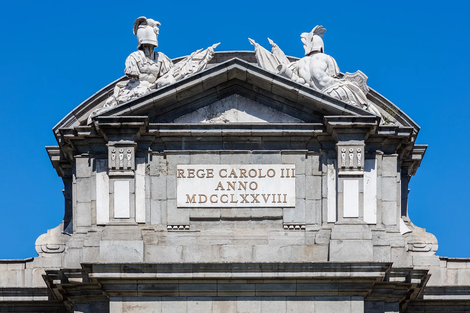 Top of the Puerta de Alcalá with two groups of carved military trophies, helmets and flags above a pediment and the plaque REGE CAROLO III ANNO MDCCLXXVIII