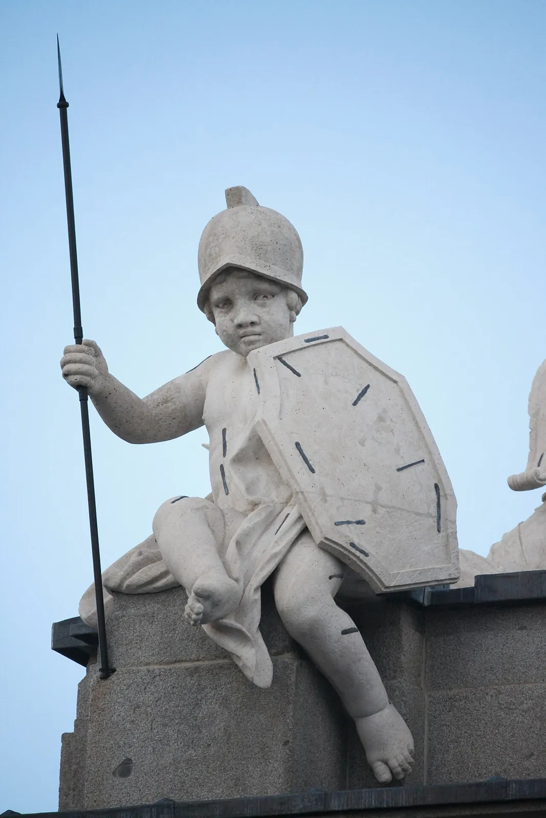 Stone statue of a seated child with a helmet, a shield and an iron spear: Fortitude, on top of the Puerta de Alcalá