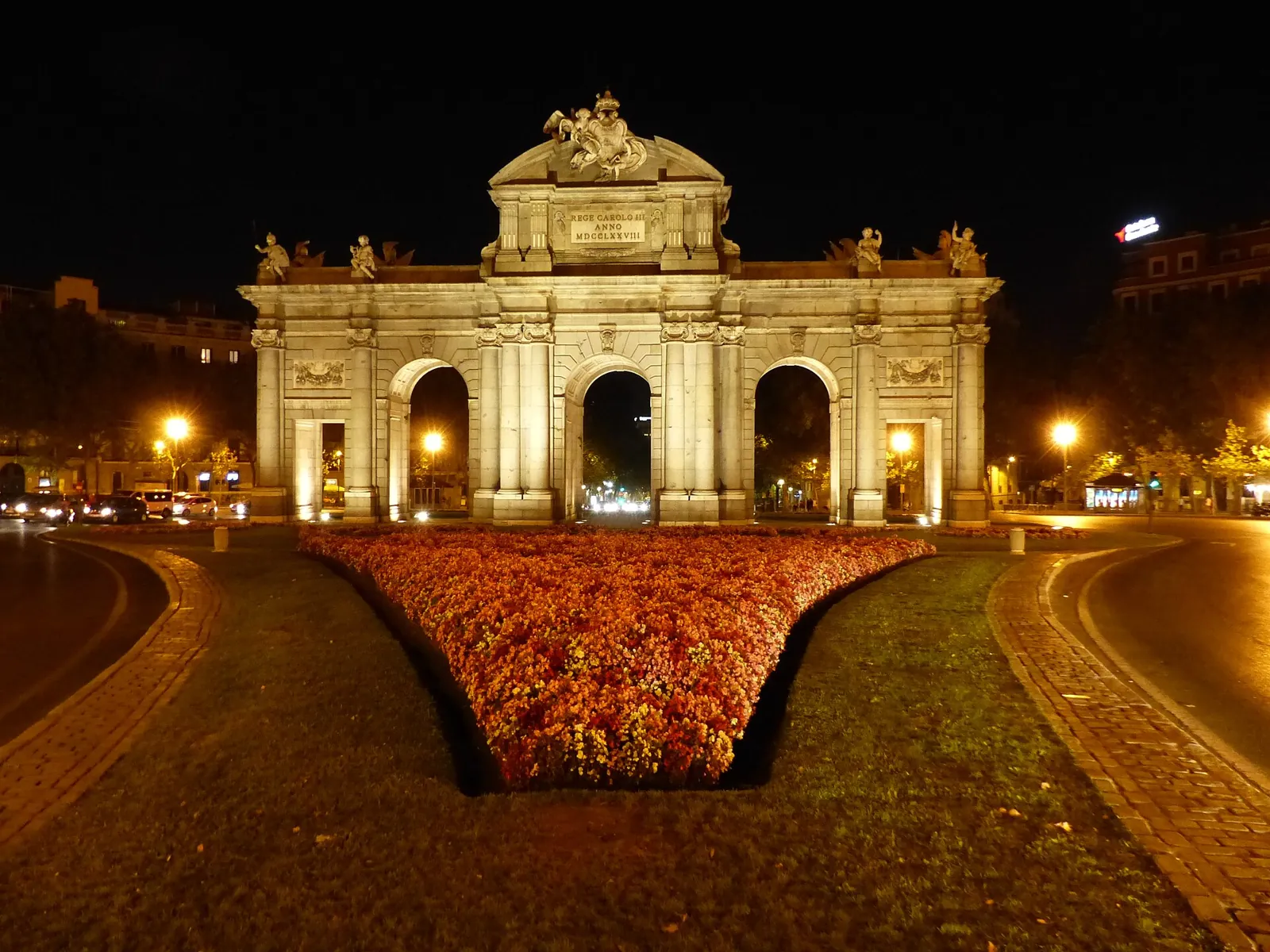 The Puerta de Alcalá lit up at night, warm light on the stone, street lamps on and a flower bed in front
