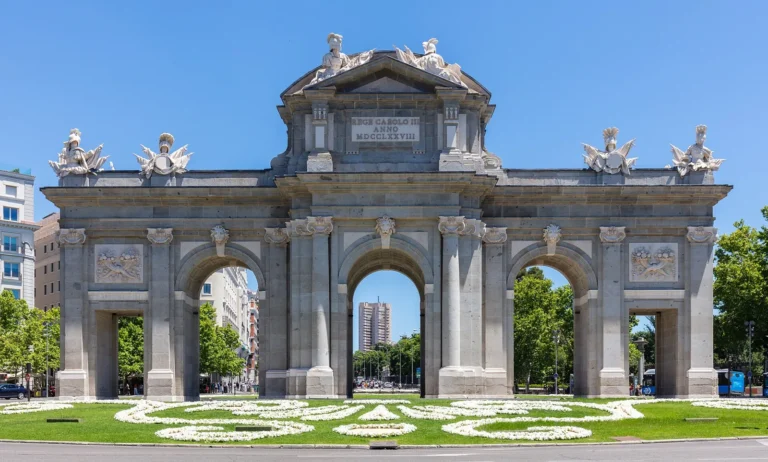 The Puerta de Alcalá seen head-on: a granite gateway with 5 openings, 3 round arches in the middle and 2 flat-topped side openings, military trophies on top and a flower bed in front