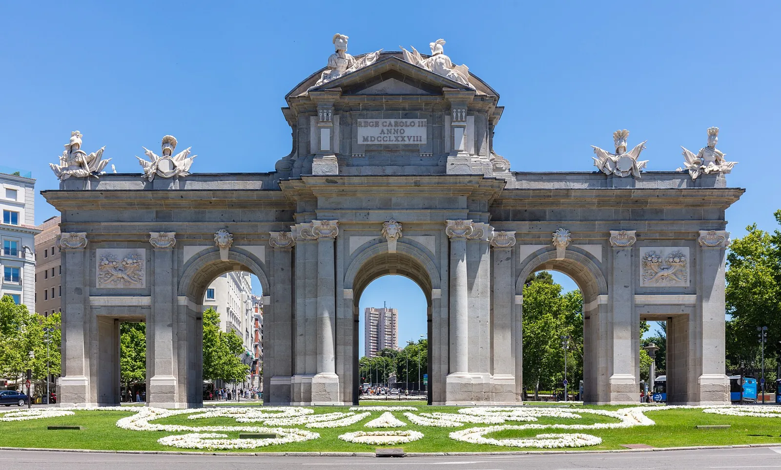 The Puerta de Alcalá seen head-on: a granite gateway with 5 openings, 3 round arches in the middle and 2 flat-topped side openings, military trophies on top and a flower bed in front