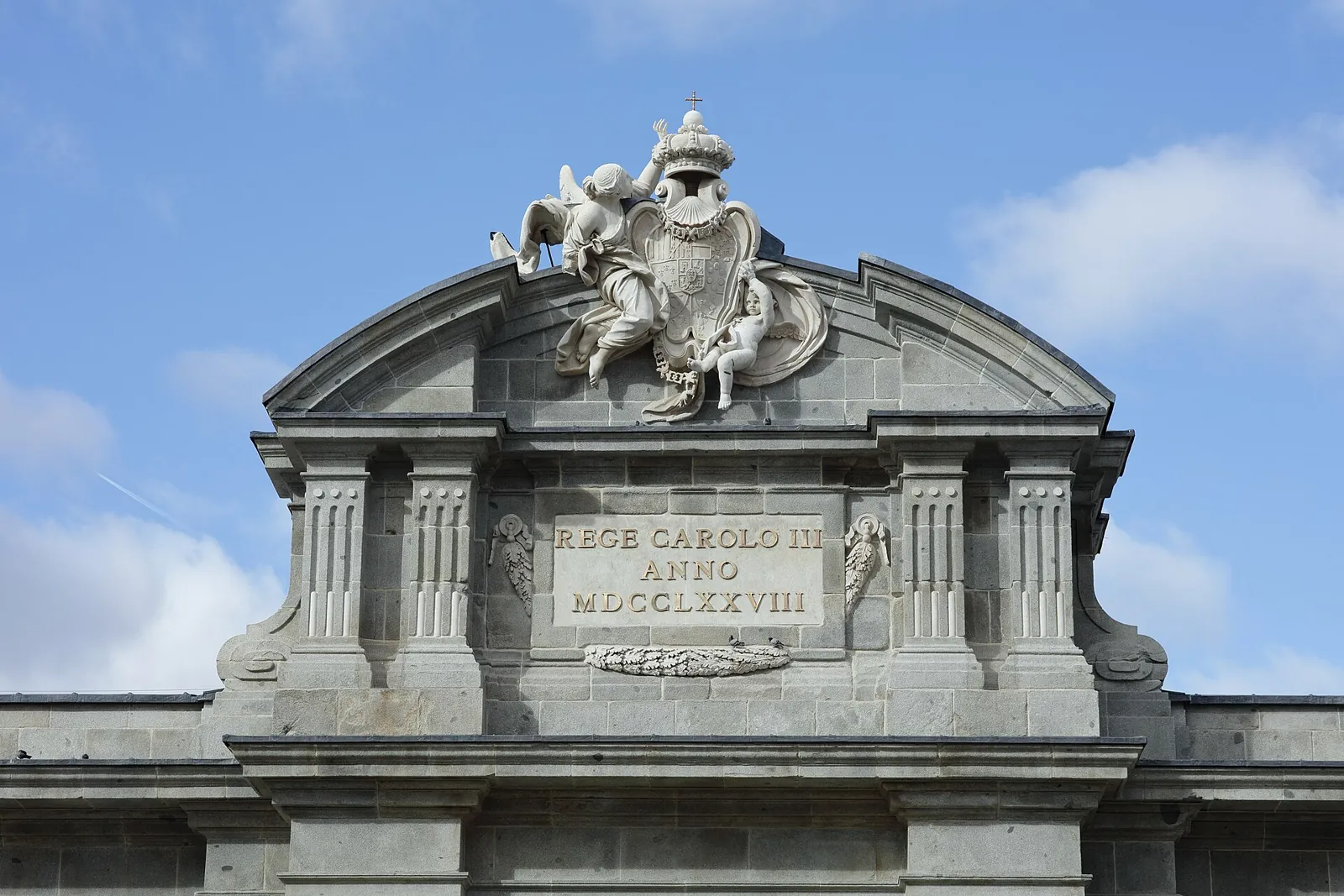 Top of the Puerta de Alcalá: the royal coat of arms held by carved figures, above a plaque reading REGE CAROLO III ANNO MDCCLXXVIII