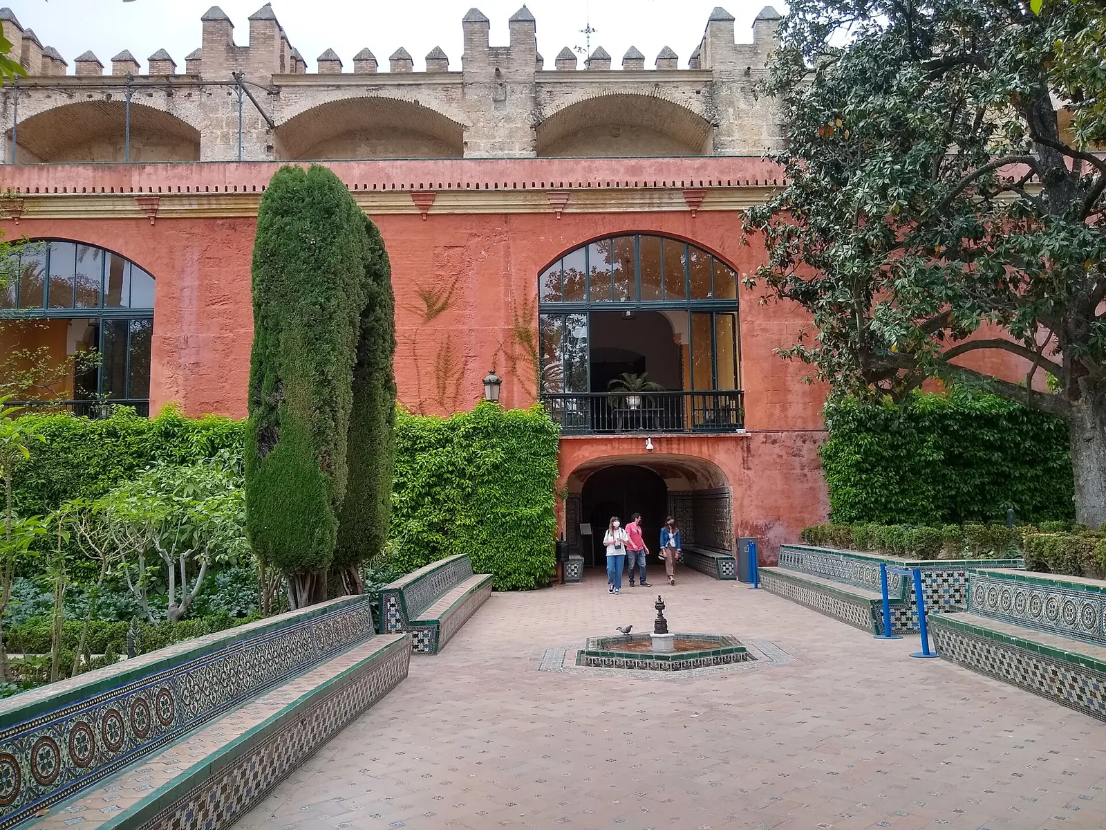 The Garden of the Dance in the Real Alcázar of Seville: a small fountain in a paved courtyard with tiled benches and hedges, in front of a red wall with a vaulted passage