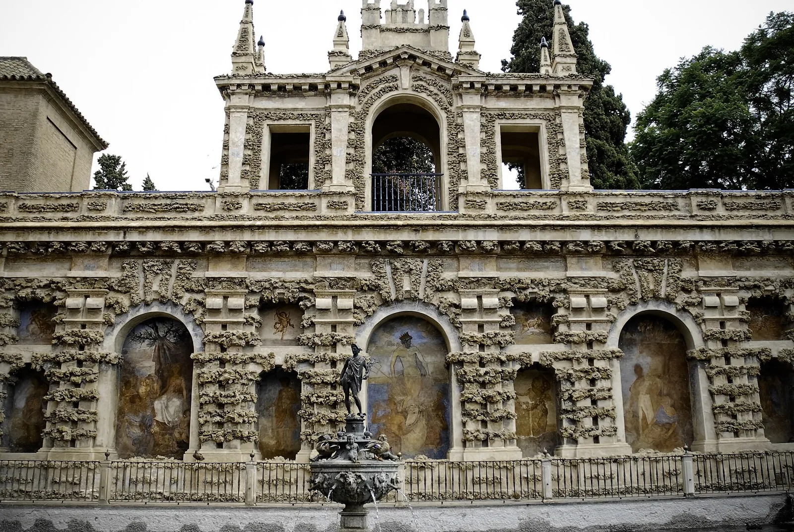 The Pond of Mercury in the gardens of the Real Alcázar of Seville: a bronze statue of Mercury on a fountain in front of a long wall of rough rustic stonework with arched niches and painted figures