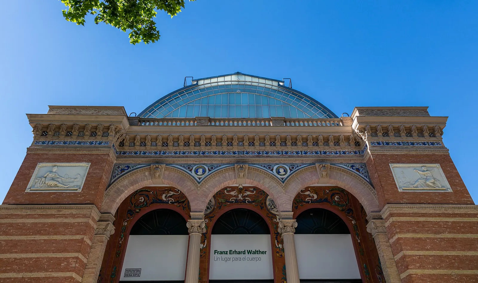 Front of the Palacio de Velázquez in Retiro Park, in two-tone brick with tiles, three entrance arches and an iron and glass vault