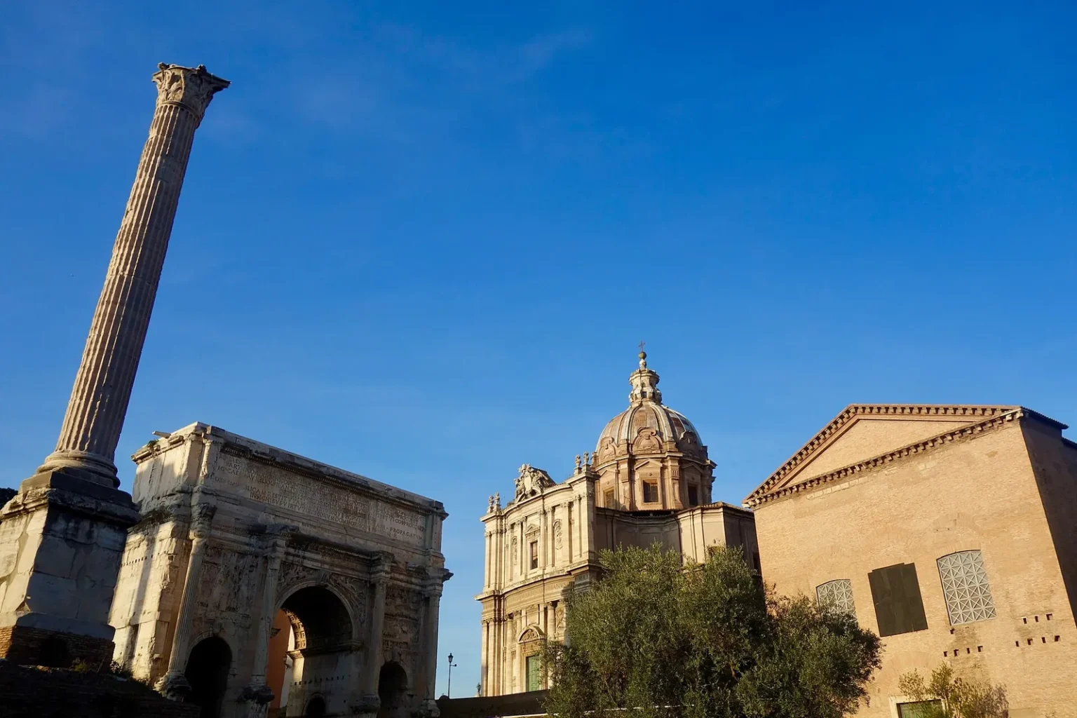 The west end of the Roman Forum against a blue sky: the tall Column of Phocas on the left, the Arch of Septimius Severus behind it, the dome of the church of Santi Luca e Martina and, on the right, the plain brick front of the Curia