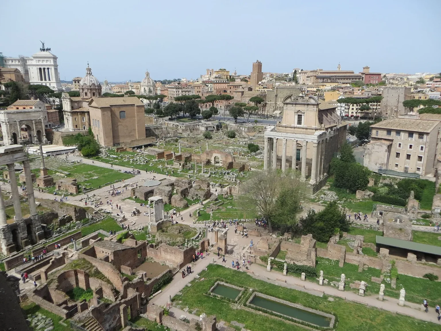 The Roman Forum seen from above on the Palatine side: the long courtyard of the House of the Vestals with its pools and statues in the foreground, the columns of the Temple of Antoninus and Faustina with a church front on the right, and the brick Curia and the Arch of Septimius Severus on the far side, under the white Vittoriano