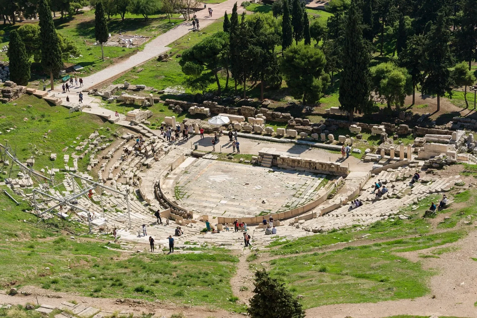 The Theatre of Dionysus seen from above: the semicircular orchestra paved in stone, the curving rows of stone seats with visitors on them, and pines and cypresses behind the stage