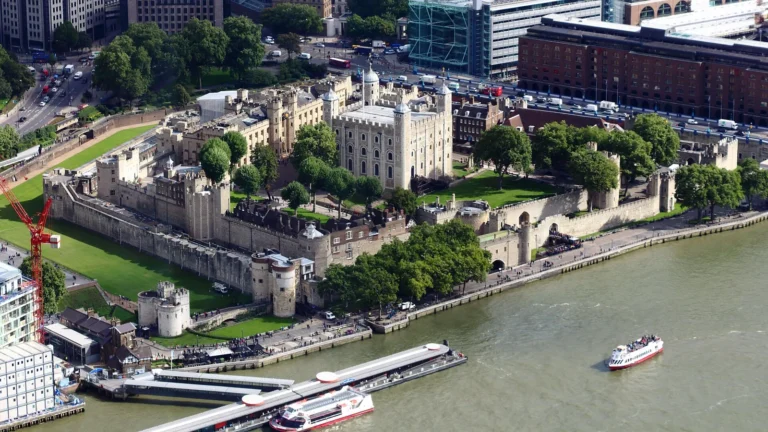 The Tower of London seen from above: the White Tower in the centre, the 2 rings of walls, the green moat and the River Thames in front with a pier and boats