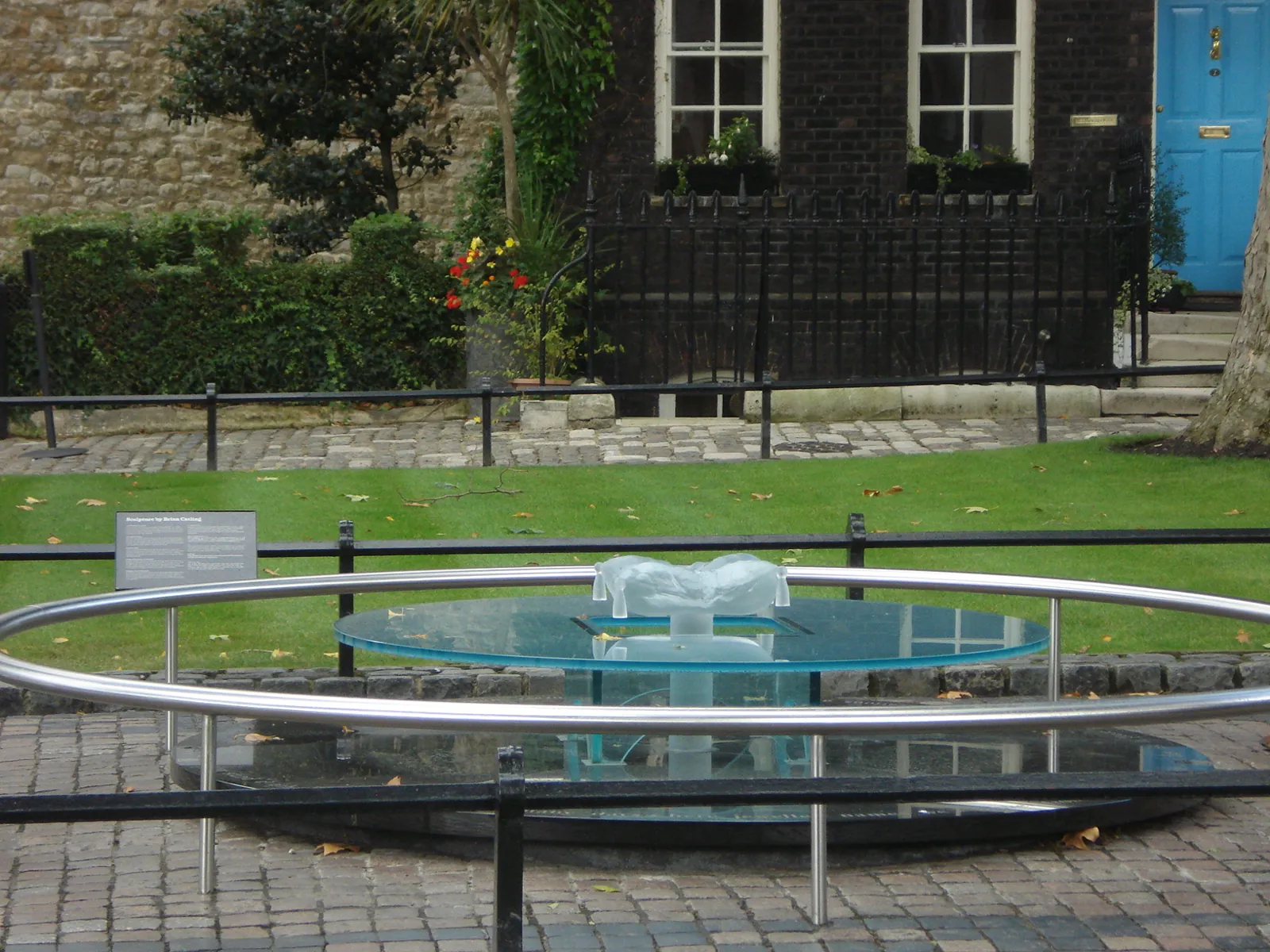The glass memorial on Tower Green: a glass disc on a low plinth inside a steel railing, with a lawn and brick houses behind