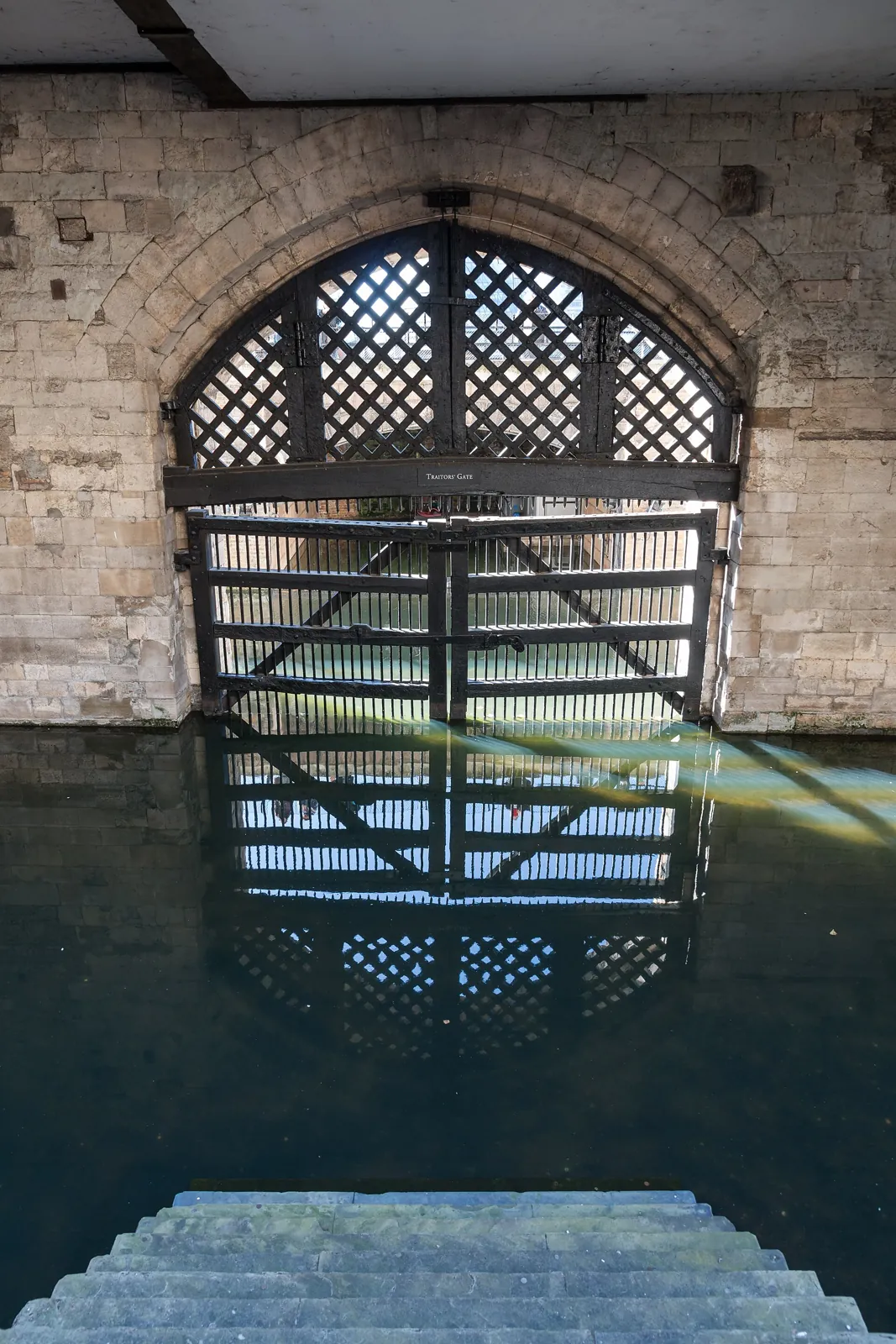 Traitors' Gate at the Tower of London: a wide stone arch closed by a heavy timber lattice gate, with dark water in front and stone steps leading down to it in the foreground
