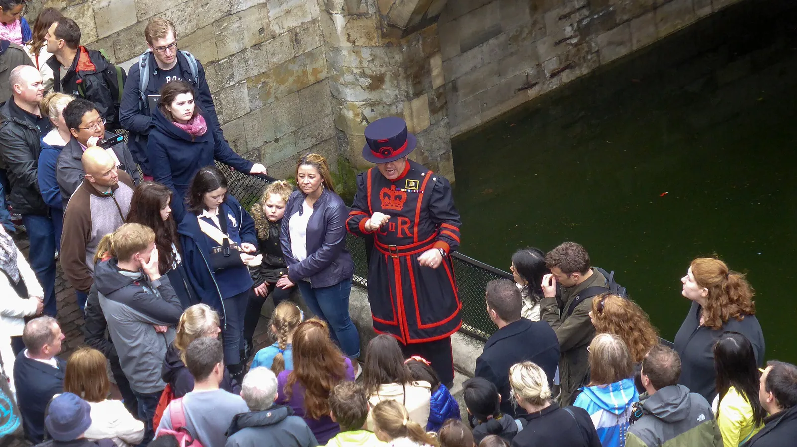 A Yeoman Warder in a dark blue and red uniform talking to a crowd of visitors beside the walls of the Tower of London