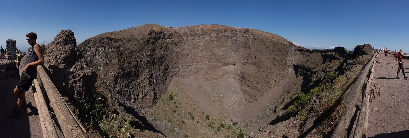 Panorama of the crater of Vesuvius from the visitor path: a wide bowl with steep walls of brown and grey layered rock, a wooden rail along the edge, a walker leaning on it on the left and people on the gravel path on the right