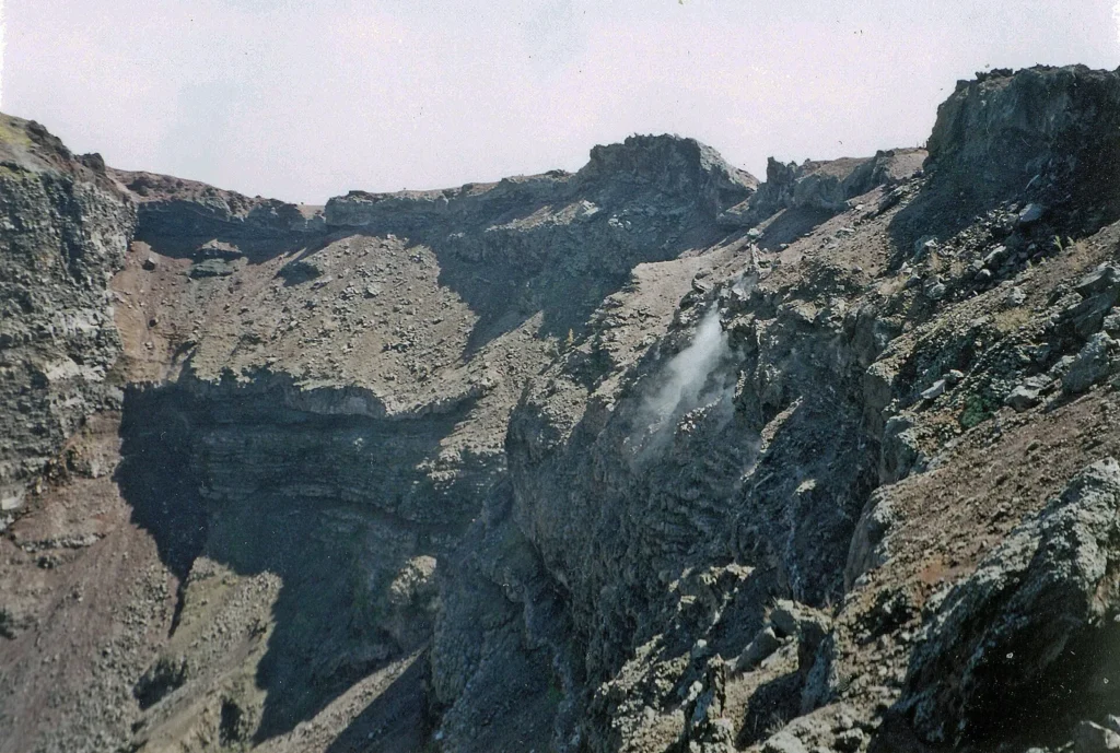 Inside the crater of Vesuvius: dark, layered walls of volcanic rock under a pale sky, with a thin plume of white steam rising from a fumarole on the right
