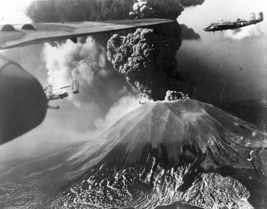 Black-and-white aerial photo of Vesuvius erupting in March 1944: a dark column of ash billowing from the summit of the cone, with American B-25 bombers flying past and the wing of another plane in the foreground